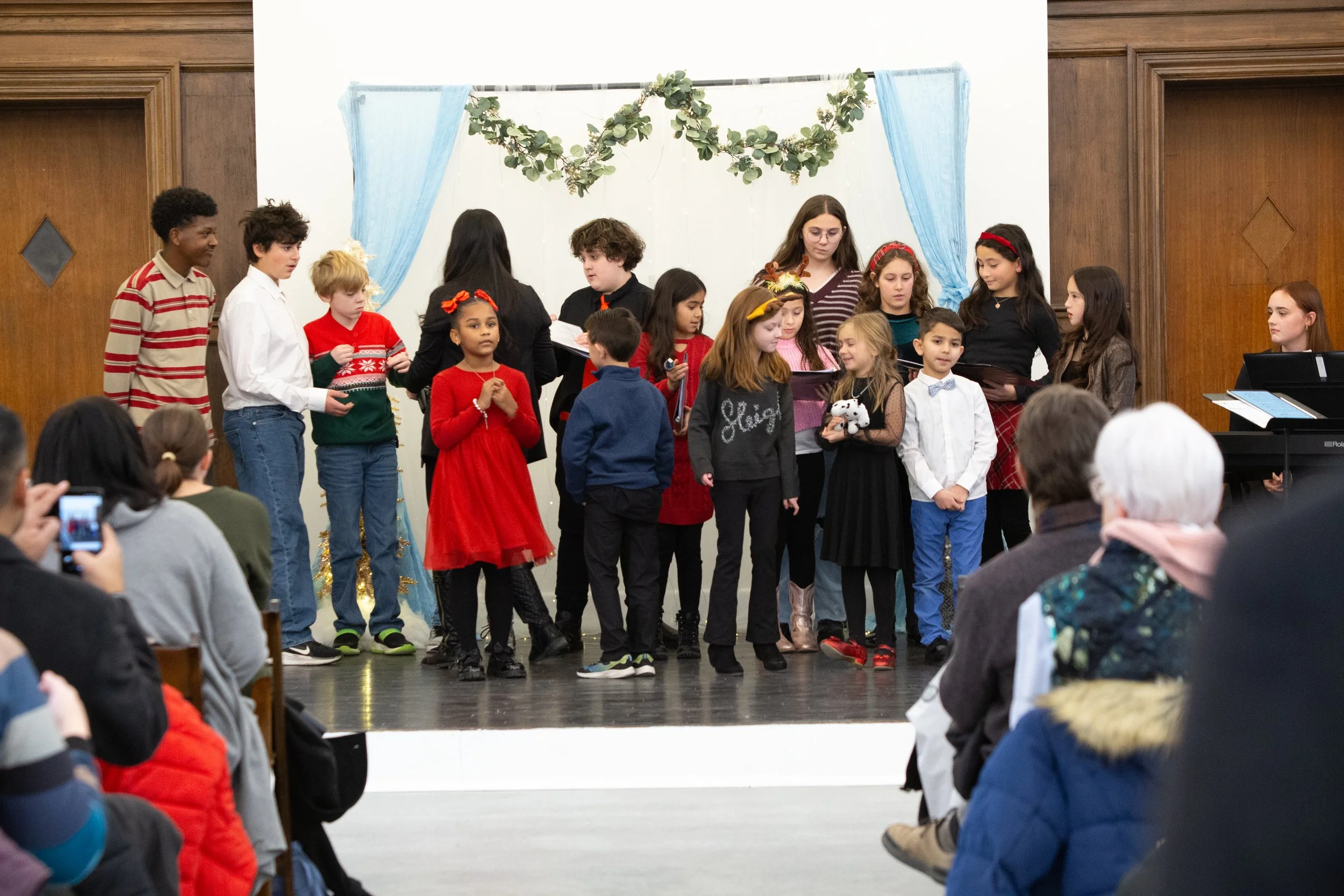 Children singing on stage during a holiday performance, with audience members watching and some taking photos.
