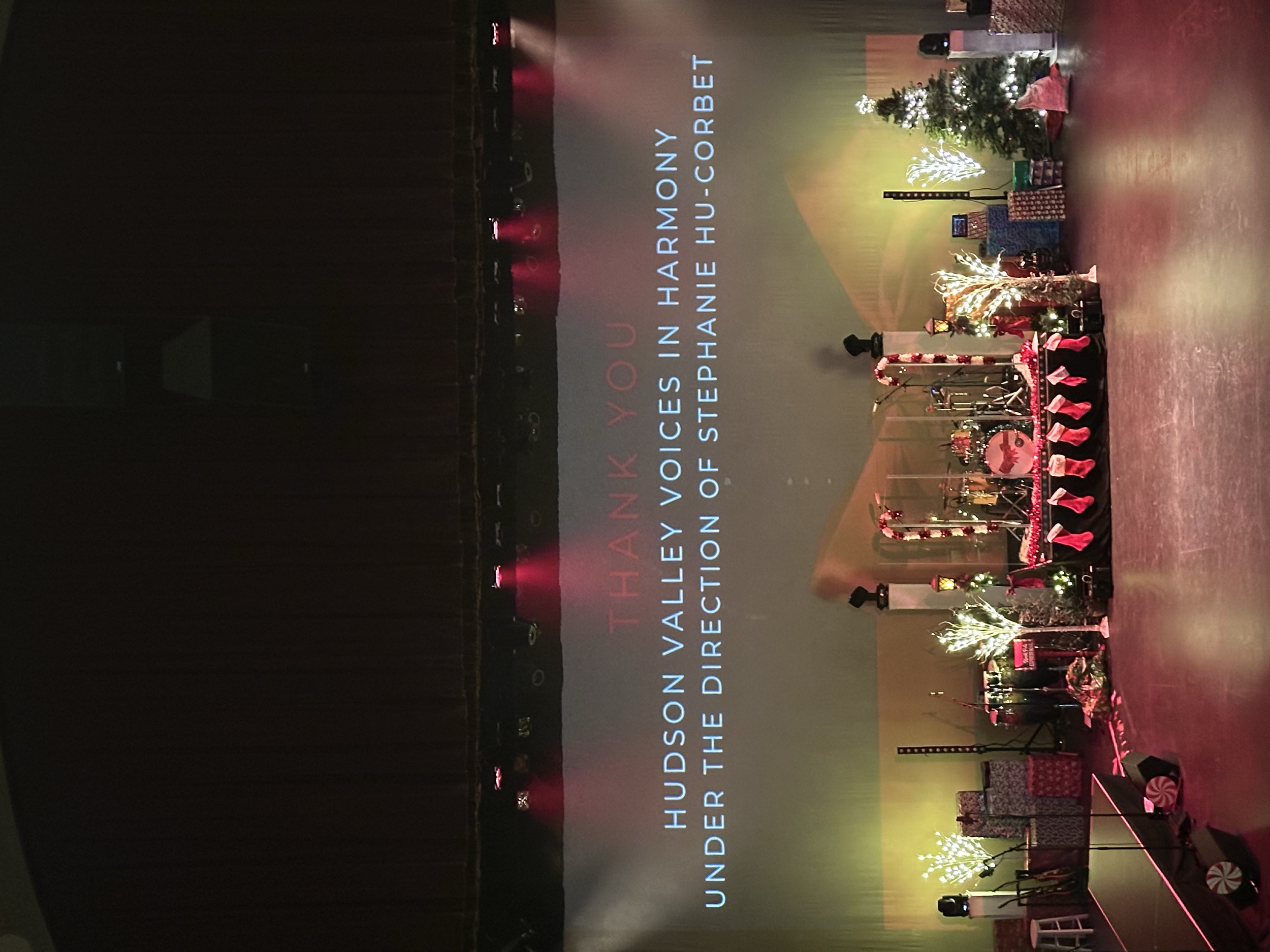 Stage decorated with Christmas trees, presents, and festive decorations with a large screen displaying a message of thanks to Hudson Valley voices in harmony under the direction of Stephanie Hu-Corbet.