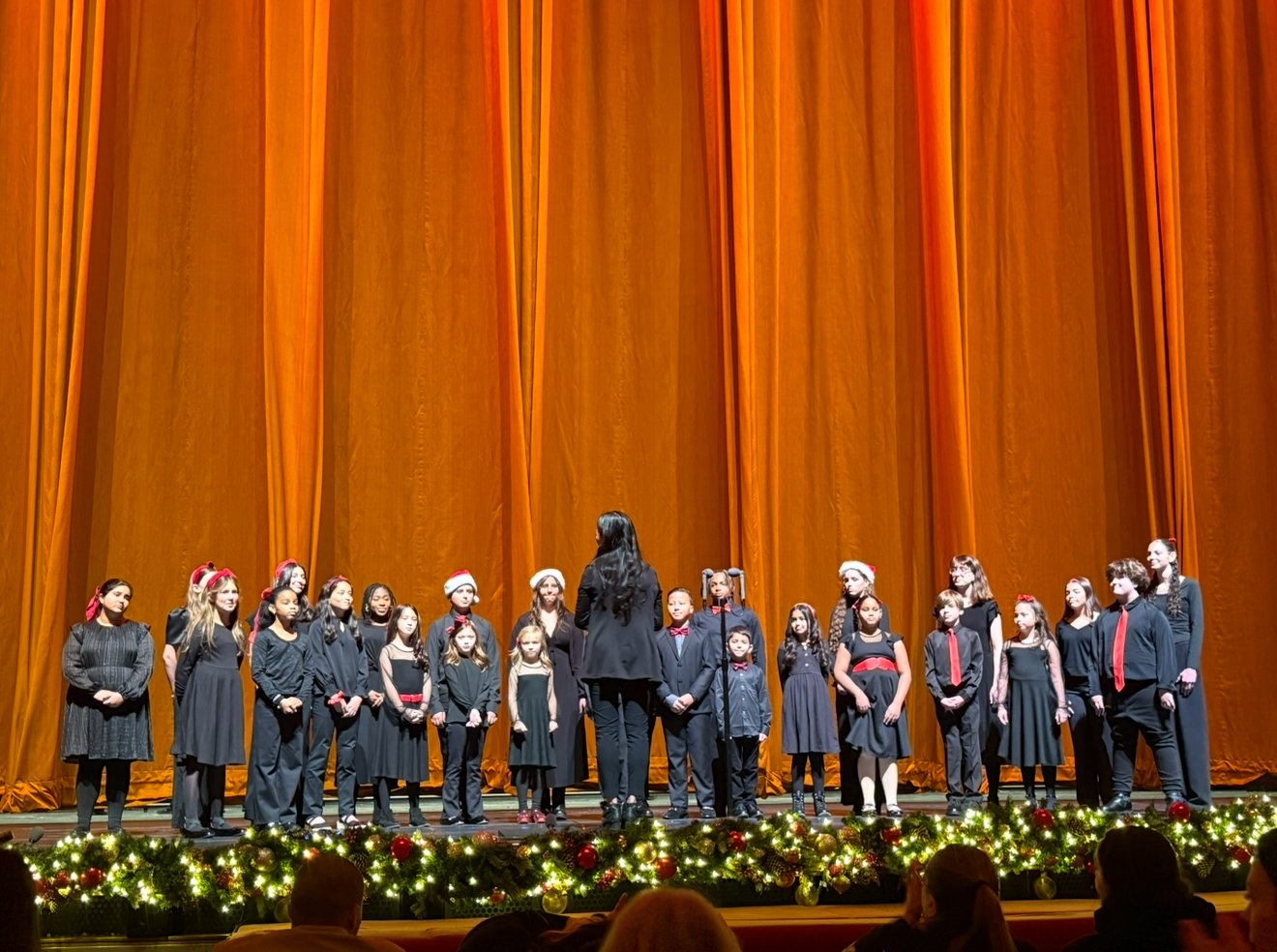 Group of children on stage with Christmas decoration, under a large orange curtain, likely performing in a holiday concert.