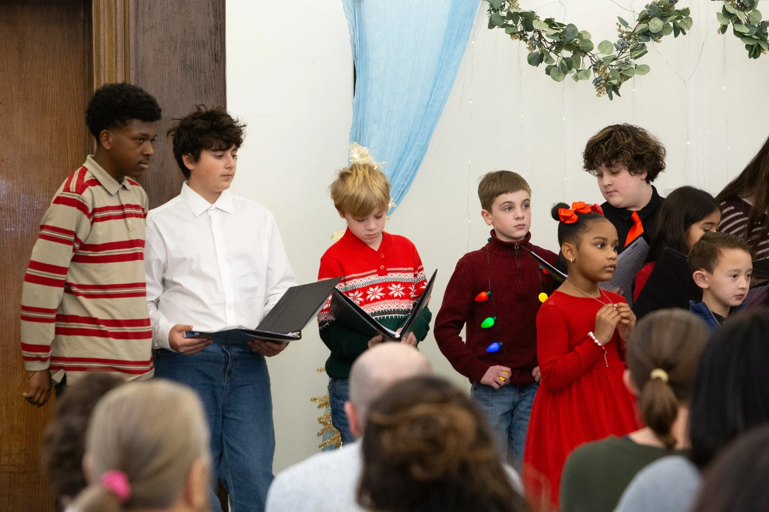 Children participating in a holiday choir performance, standing in front of an audience, dressed in festive holiday attire, with Christmas decorations in the background.
