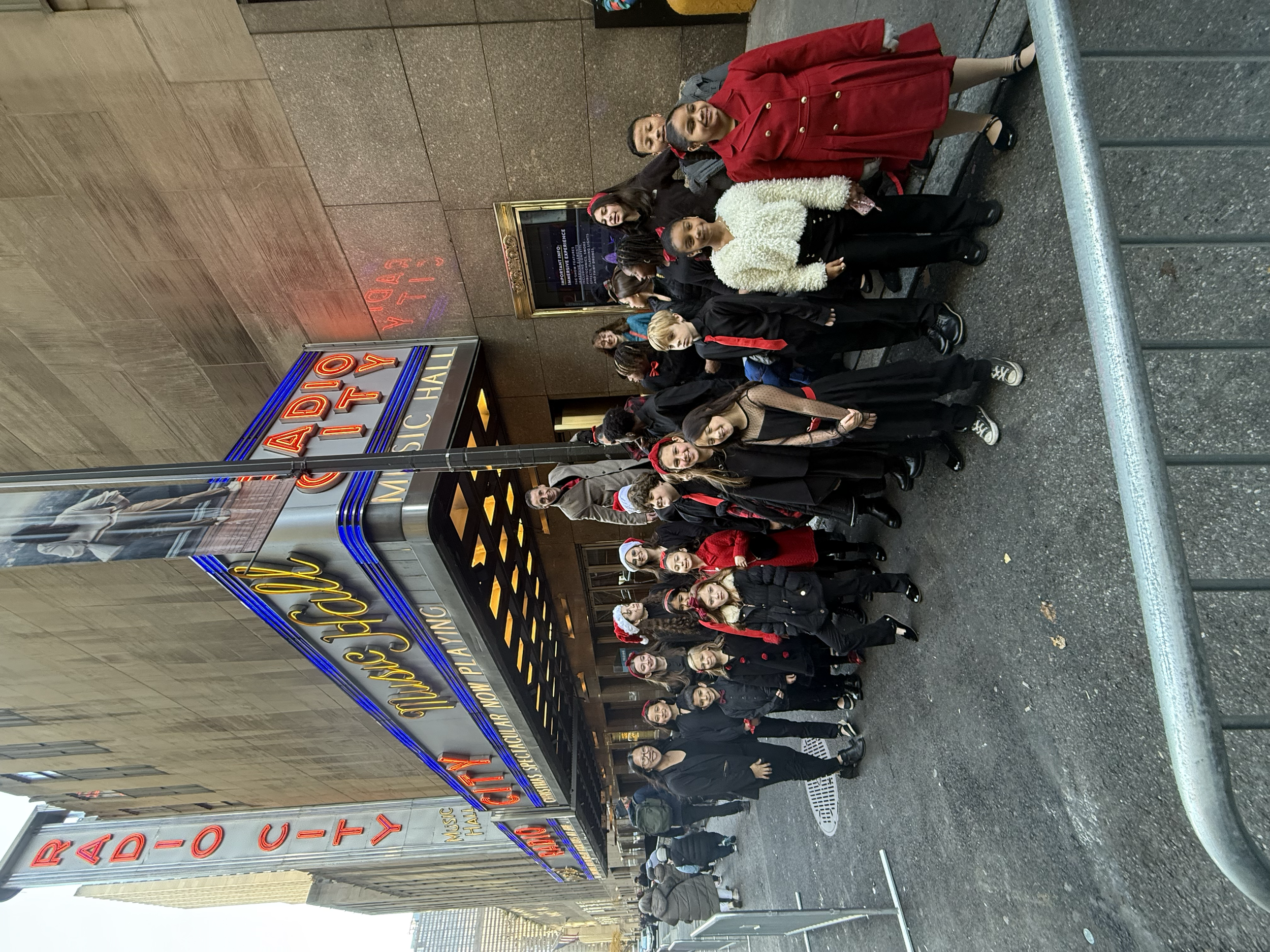 A group of people is standing in front of the Radio City Music Hall marquee, dressed in black and red, some wearing Santa hats. They appear to be posing for a photo outside the theater.