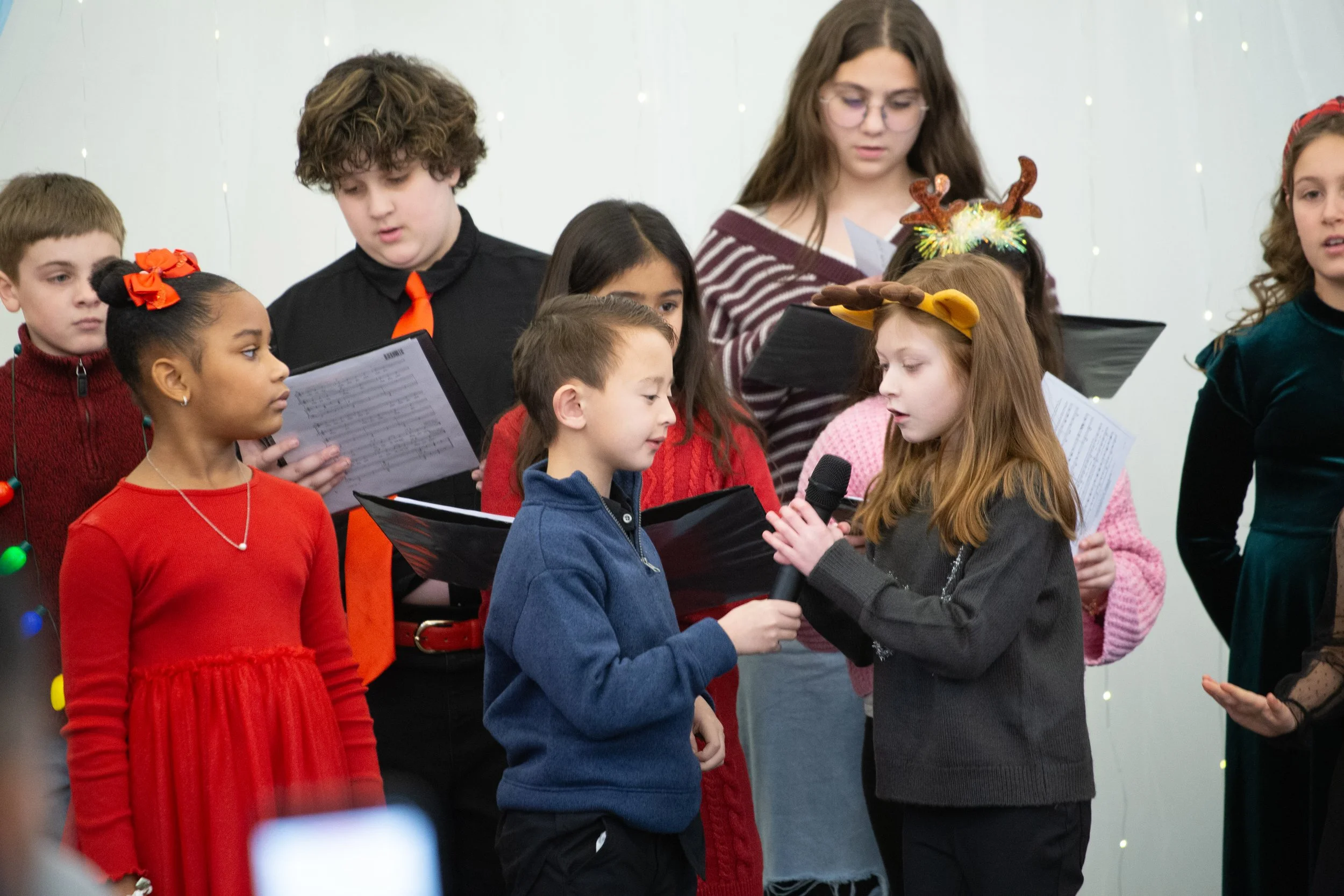 Children dressed in festive clothing, some with reindeer antlers and Christmas accessories, singing and holding a microphone during a Christmas performance or choir event.