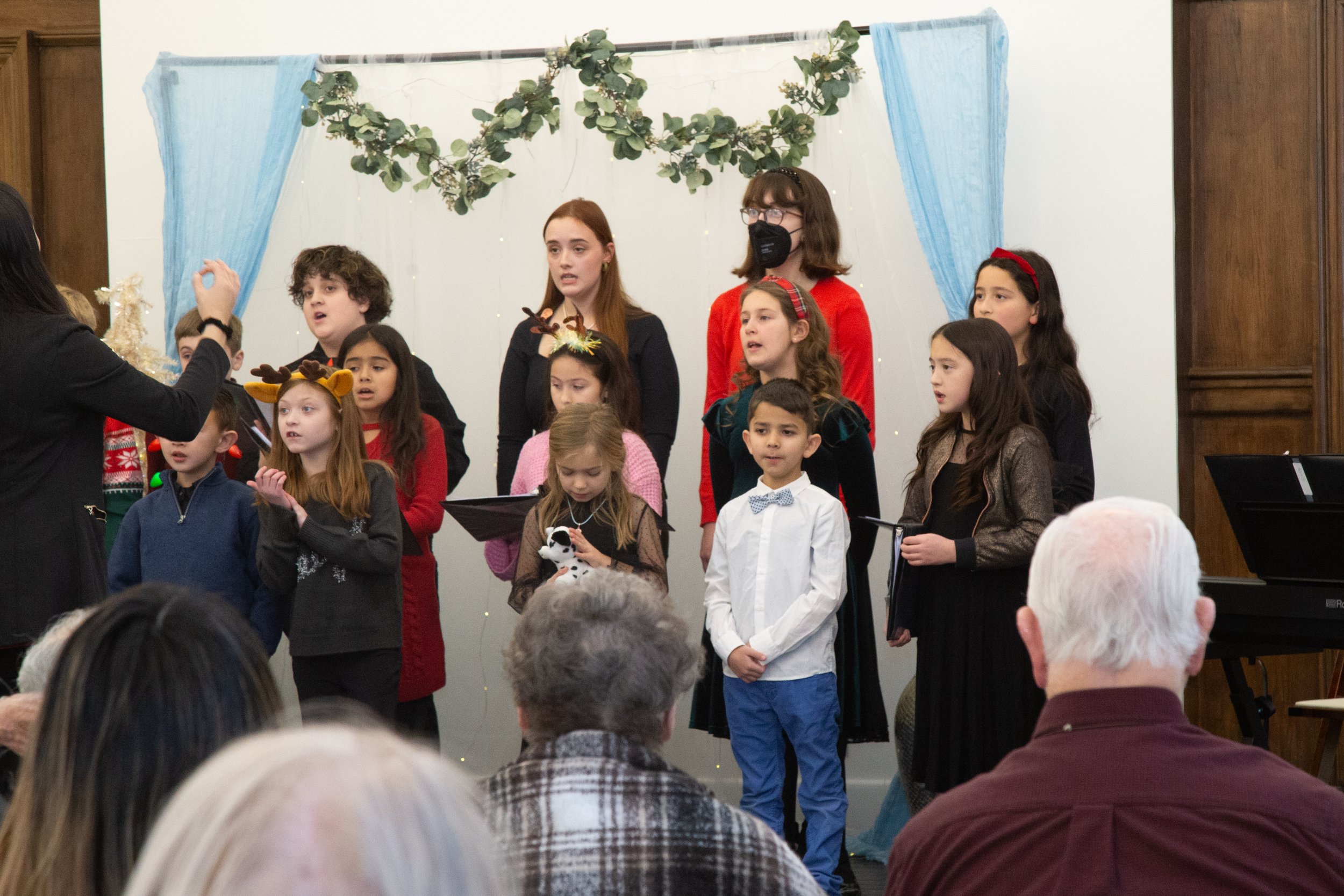 Children's choir performing on stage with a Christmas-themed backdrop, some children wearing holiday accessories like reindeer antlers and holding songbooks, during a holiday event.