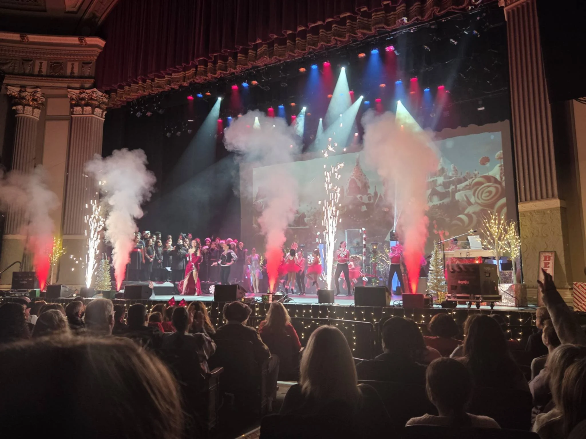 Stage with performers, colorful lights, fireworks, and Christmas decorations during a holiday show, with audience watching.