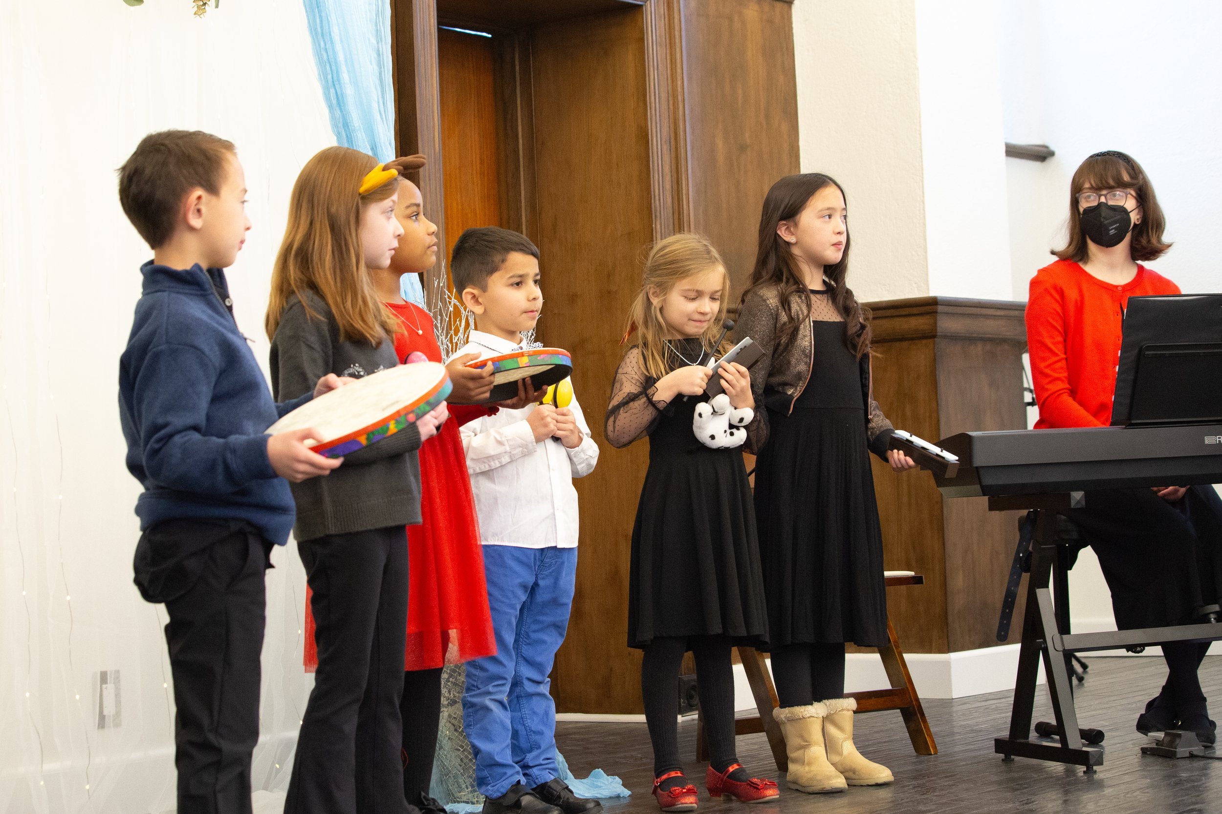 Children performing at a school concert, standing in a line with some holding percussion instruments, and a woman playing a keyboard, all indoors.