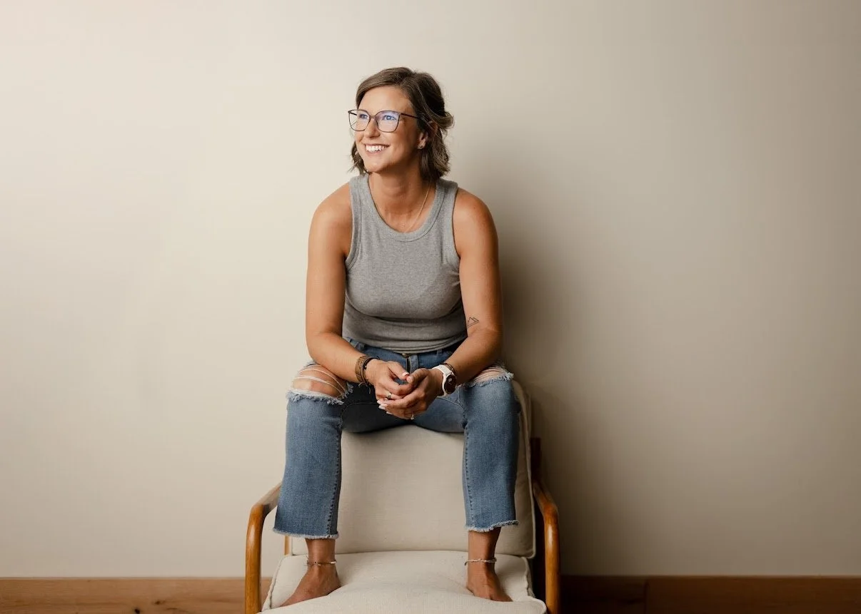 A woman with glasses, wearing a gray sleeveless top and ripped jeans, sitting on a white chair against a plain beige wall, smiling and looking to the left.