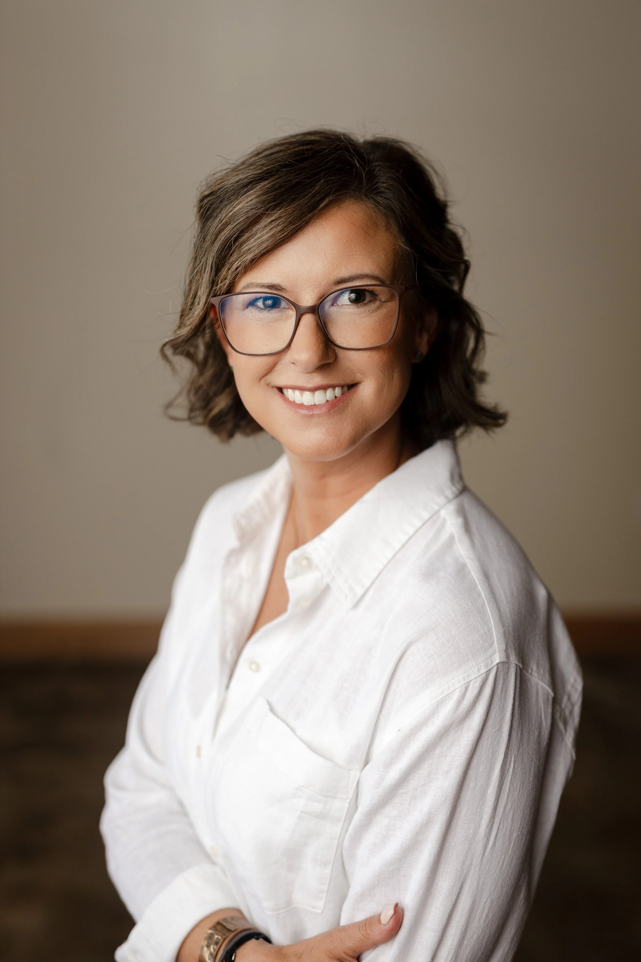 A woman with short brown hair, glasses, and a white shirt is smiling at the camera.