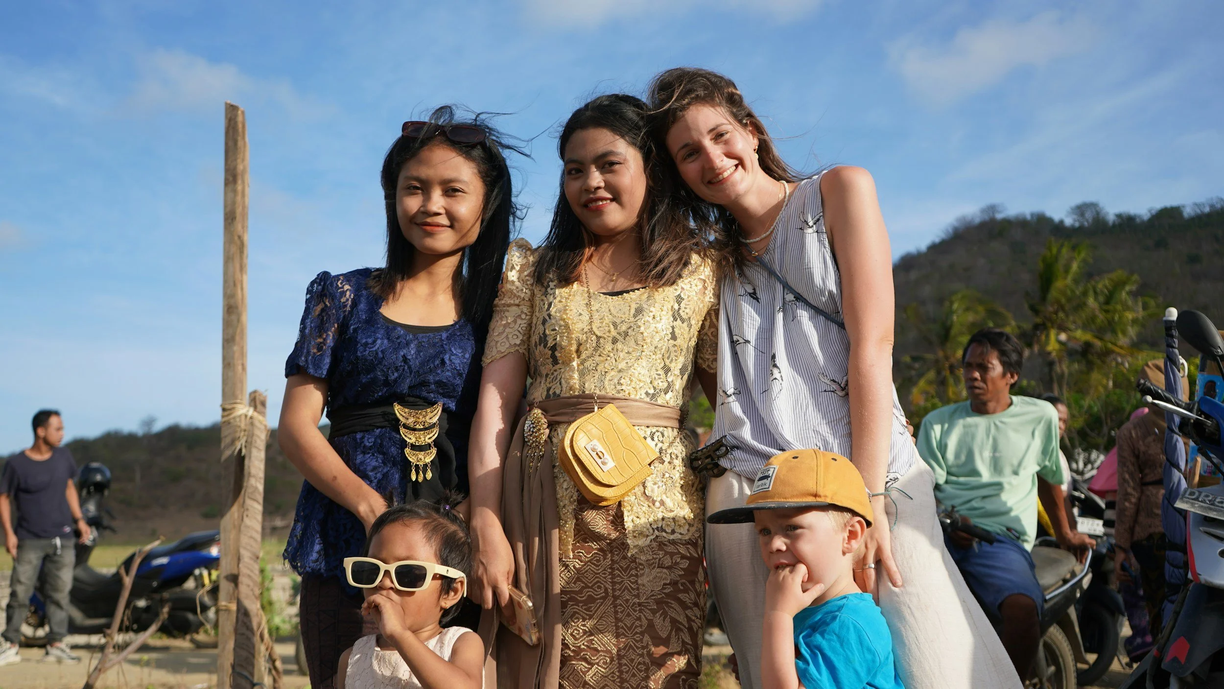 A group of women and children standing outdoors in a sunny area, with motorcycles and other people in the background, some on a dirt road or field, with hills and trees visible under a blue sky.