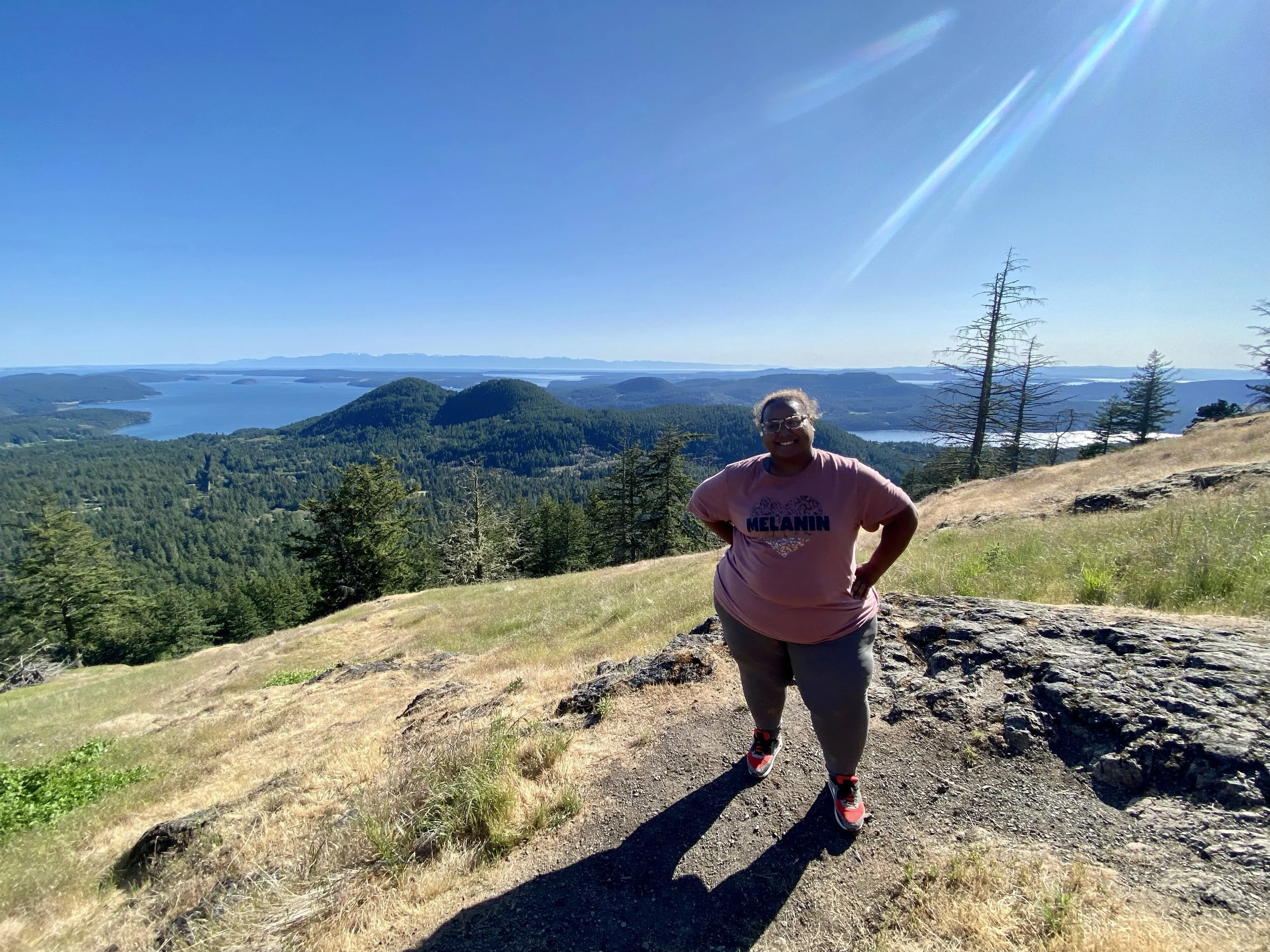 A person in a pink shirt with 'MELANIN' written on it, smiling and standing on a trail on a hilltop overlooking a landscape of lakes, forested hills, and mountains under a clear blue sky with sunlight shining.