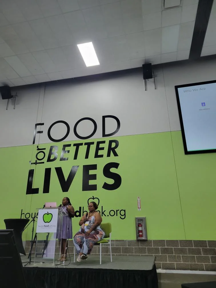 Two women talking on a stage in front of a large green and white wall with the words 'Food for Better Lives' and a website address. One woman is standing with a microphone, and the other is sitting on a chair.