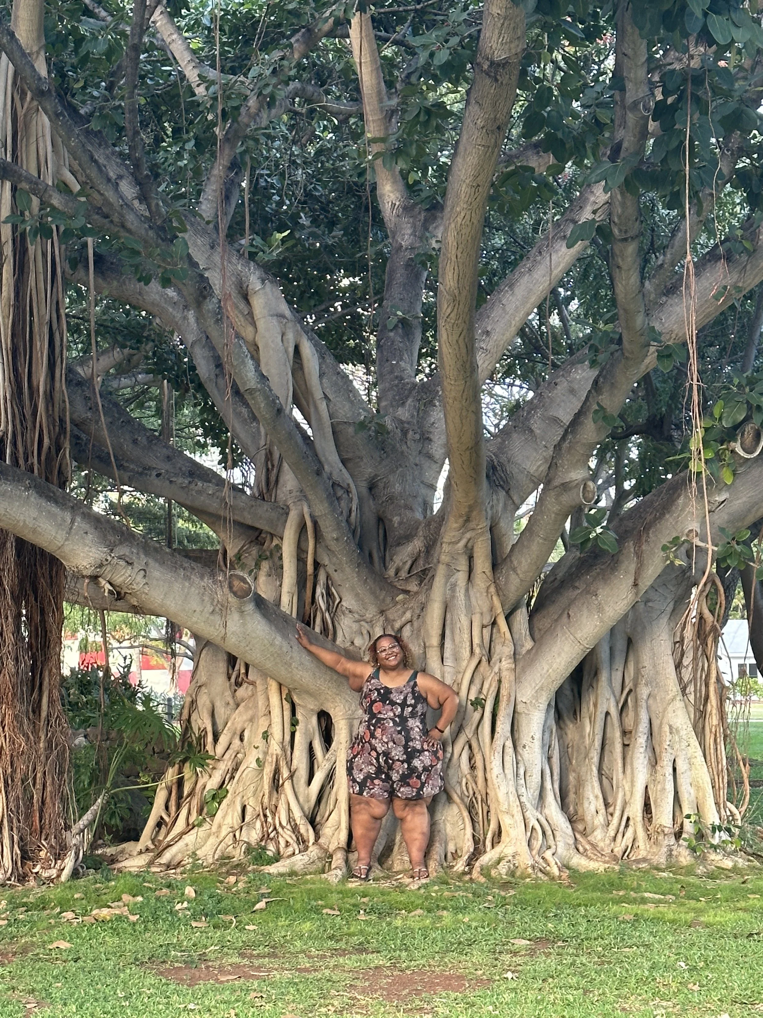 A woman in a floral dress standing in front of a large tree with numerous thick roots and branches. She is smiling and posing, with one arm resting on a tree branch.