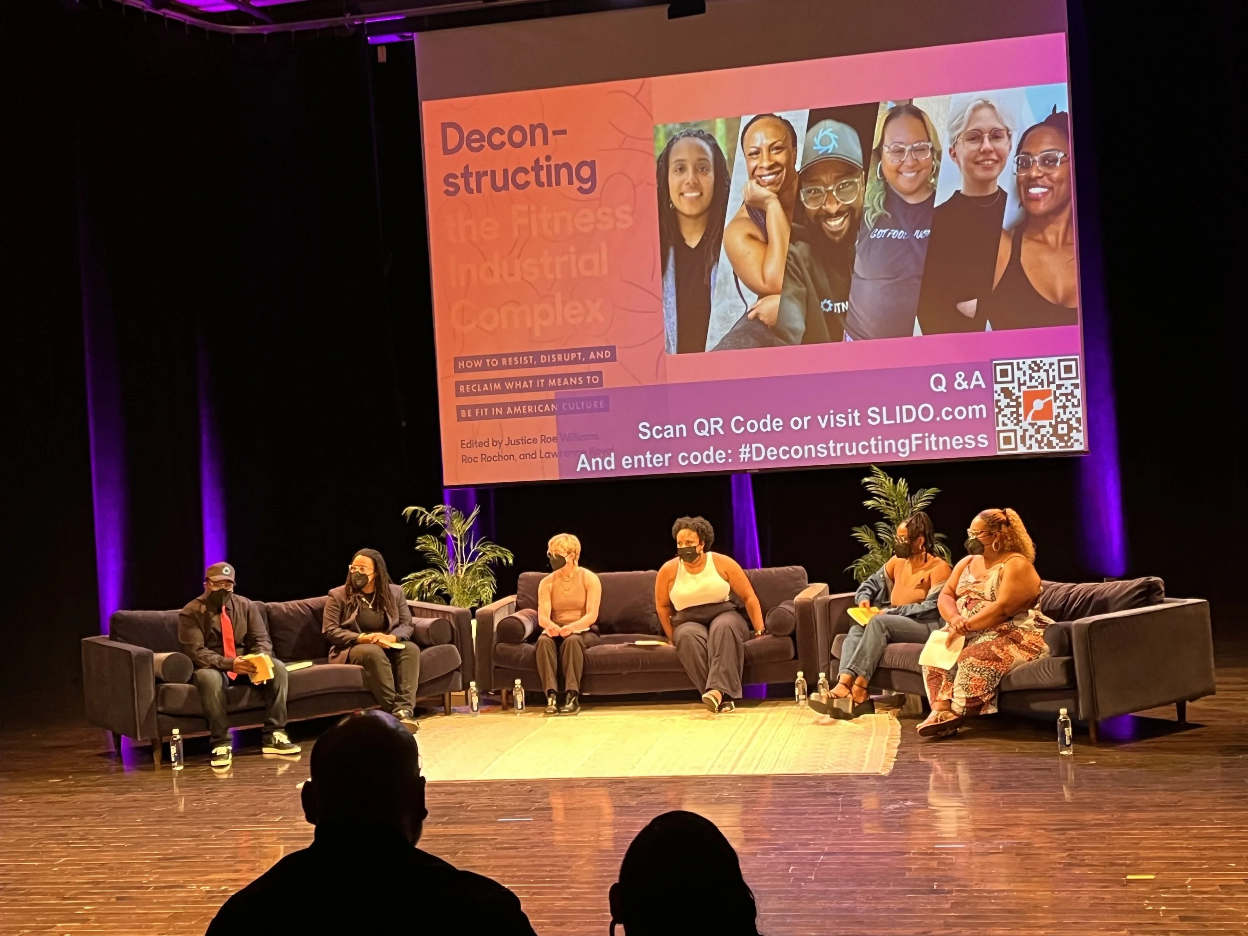 A panel discussion with six women seated on a stage with black sofas, facing the audience. Behind them, a large screen displays a colorful presentation slide about deconstructing the fitness industrial complex, featuring a group photo of diverse women, a QR code, and text instructions for accessing additional information.