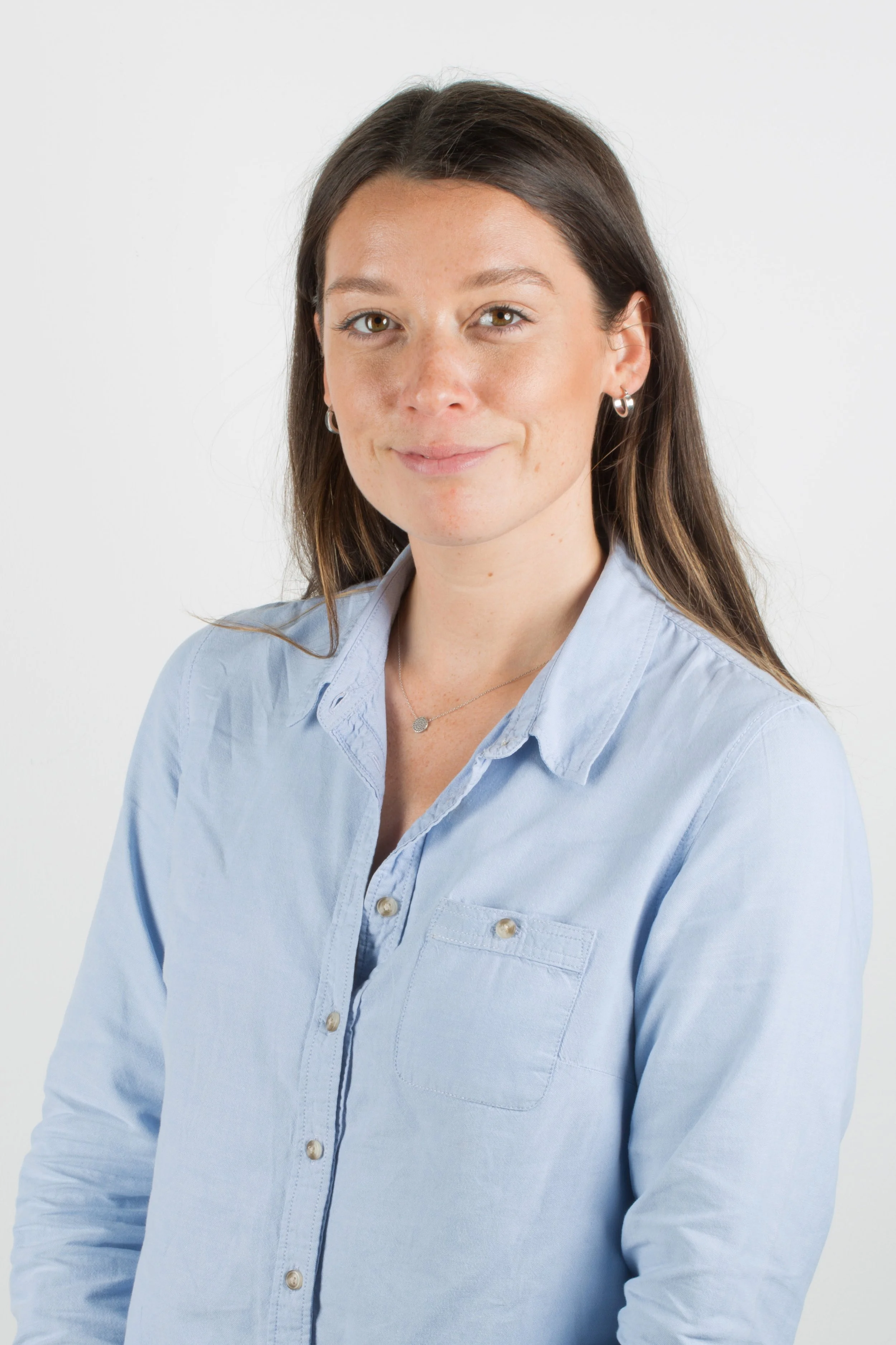 A woman with long brown hair and light skin, wearing a light blue button-up shirt, earrings, and a necklace, smiling at the camera against a plain white background.