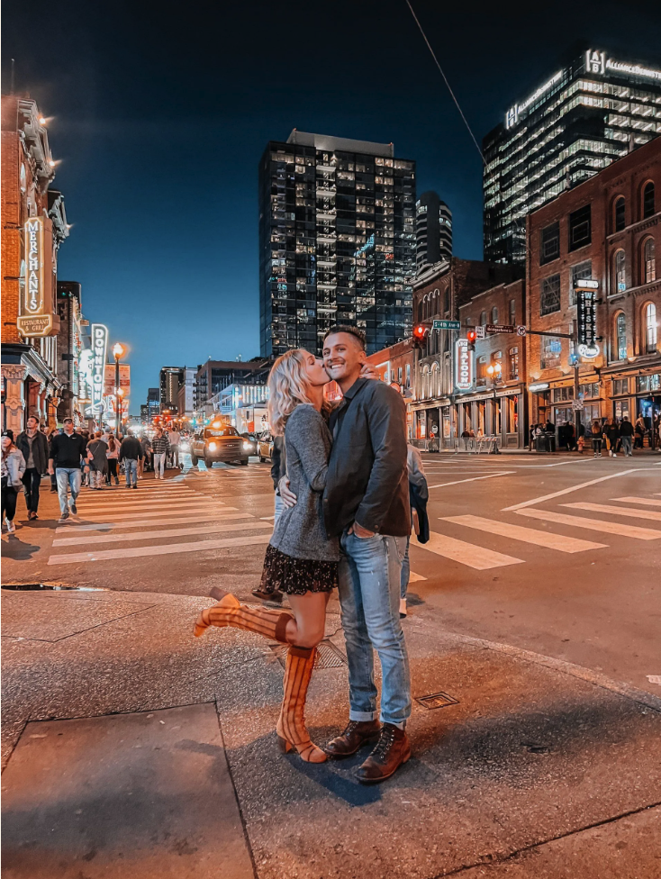 A young couple sharing a kiss and a hug on a city street at night, with illuminated buildings and pedestrians in the background.