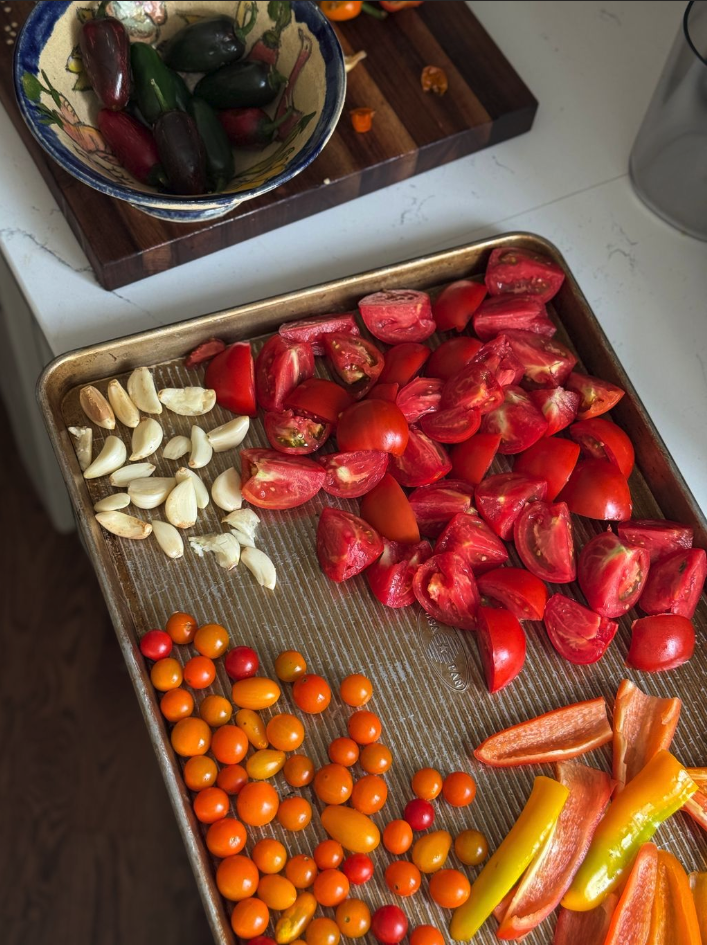 Assorted fresh vegetables on a metal tray and a wooden cutting board, including cherry tomatoes, halved tomatoes, garlic cloves, and sliced peppers.