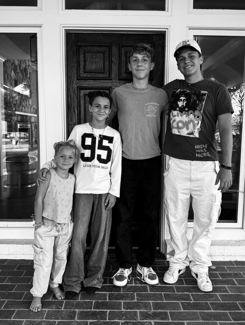 Five children standing on a porch in front of a wooden door, posing for a photo. The group includes two young girls and three boys, smiling at the camera.