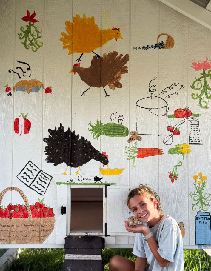 Young girl smiling and holding a small bird in front of a painted mural of chickens, vegetables, farm produce, and farm-related items on a white wall.