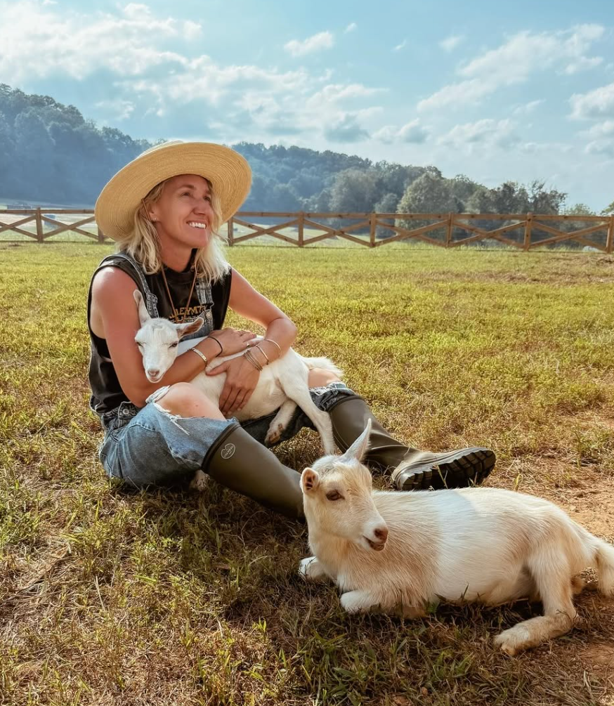 A woman wearing a large straw hat, black sleeveless top, ripped jeans, and rain boots sits on grass in a rural setting, holding a small white goat. There is another white goat lying on the ground beside her. In the background, there is a wooden fence