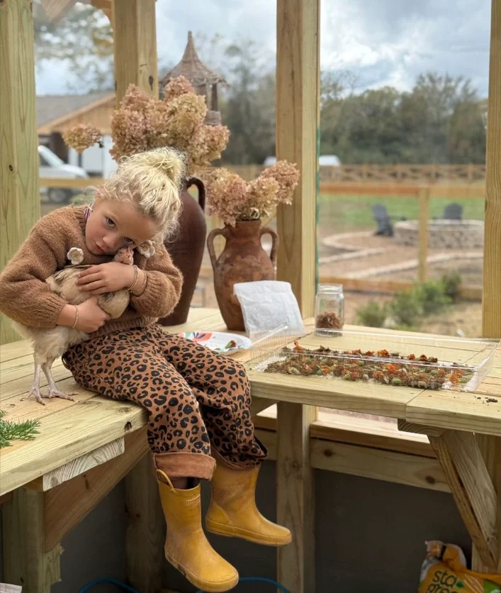 A young girl with curly blonde hair sitting on a wooden bench holding a chicken, wearing leopard print pants and yellow boots, in a rustic outdoor space with potted plants and blurred background.