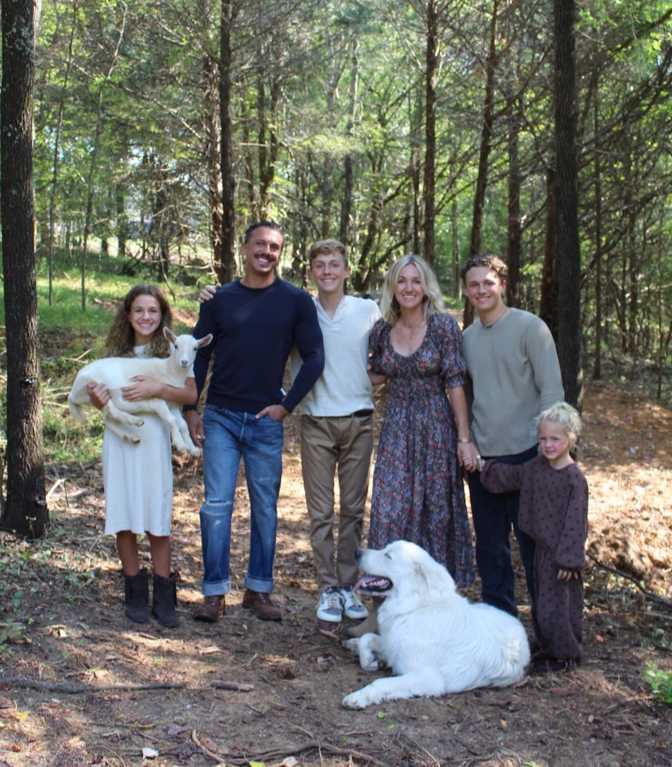 A family of seven, including two children, two adults, two dogs, and a young goat, standing in a wooded forest with sunlight filtering through the trees.