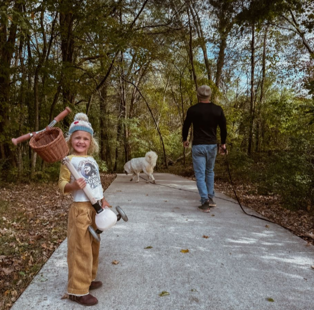 A young girl smiling, holding a basket and a toy, standing on a sidewalk in a wooded park, with a man walking a dog in the background.