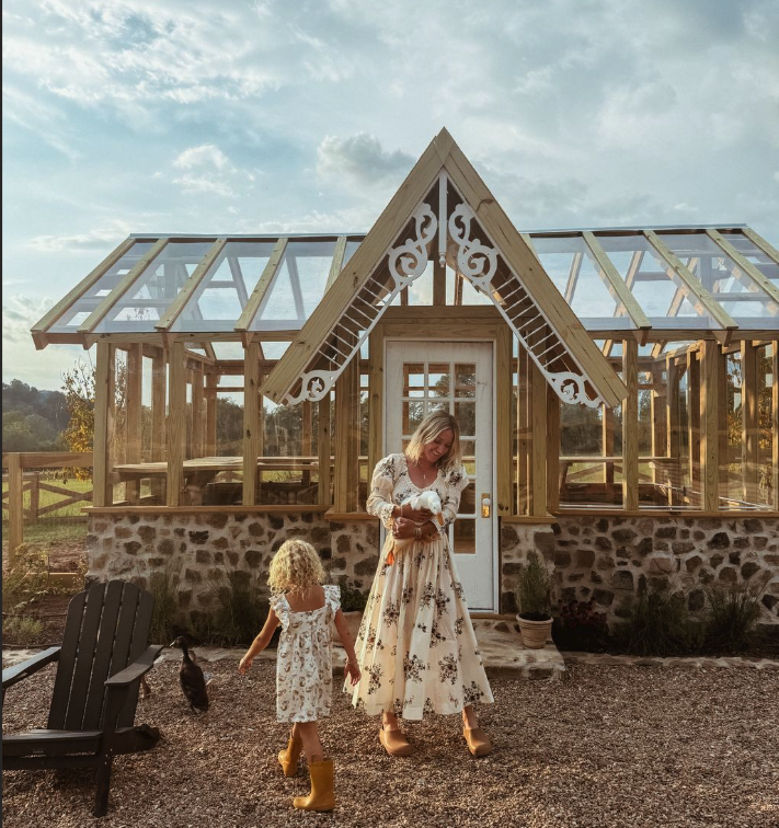 A woman and a young girl standing outside a glass greenhouse with decorative wood trim. The woman is holding a small dog, and the girl is walking towards a black cat. The scene is set on a sunny day with a partly cloudy sky.