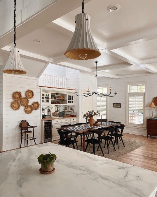 Dining area with a black table, six black chairs, a chandelier, and natural light from several windows in a white room.