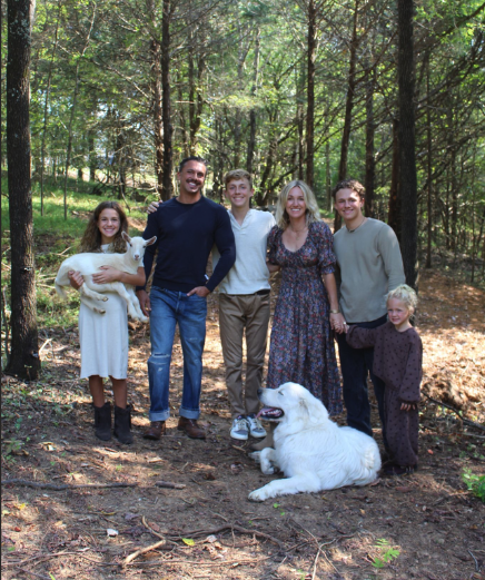 Family of six with one dog standing in a forest clearing on a sunny day.