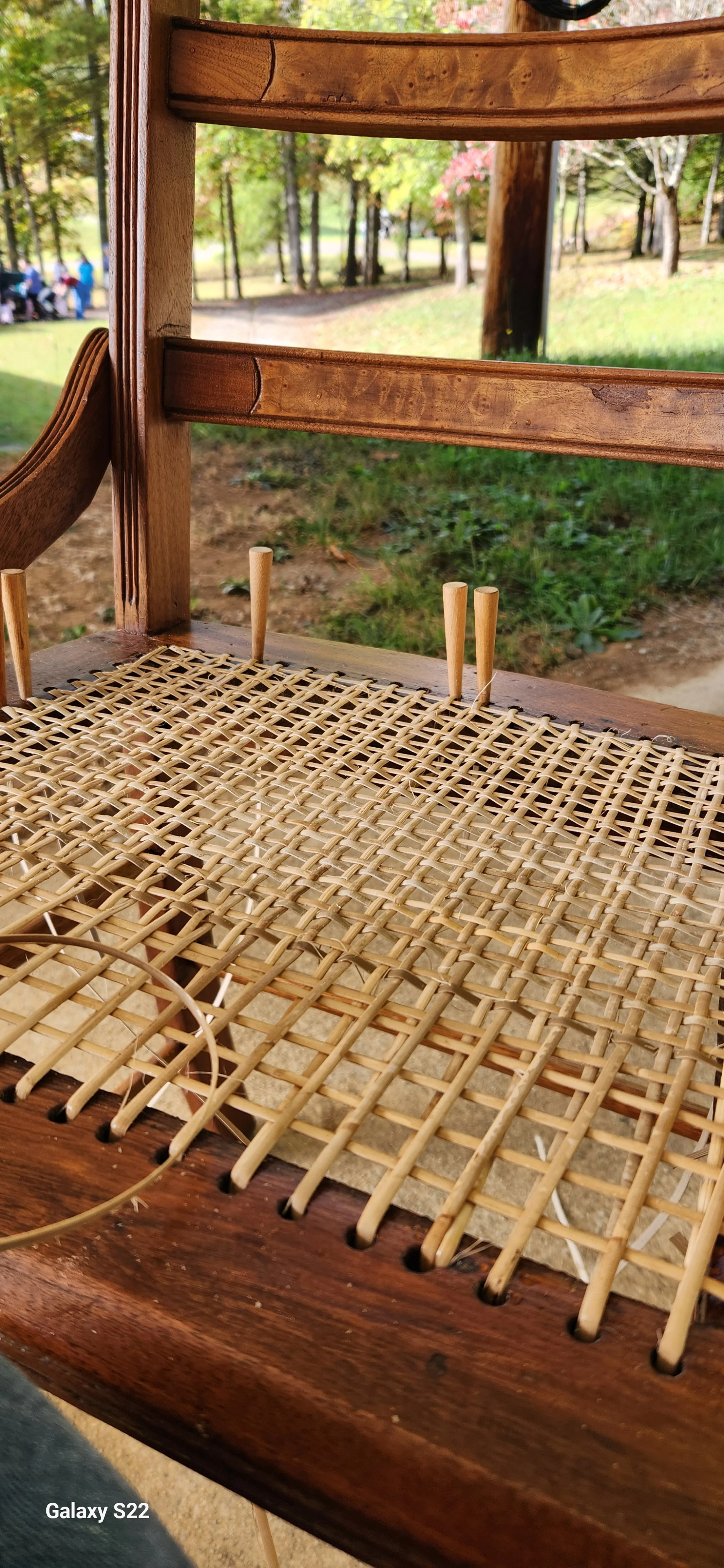 A wooden chair with a woven cane seat, outdoors in a park setting with trees in the background.