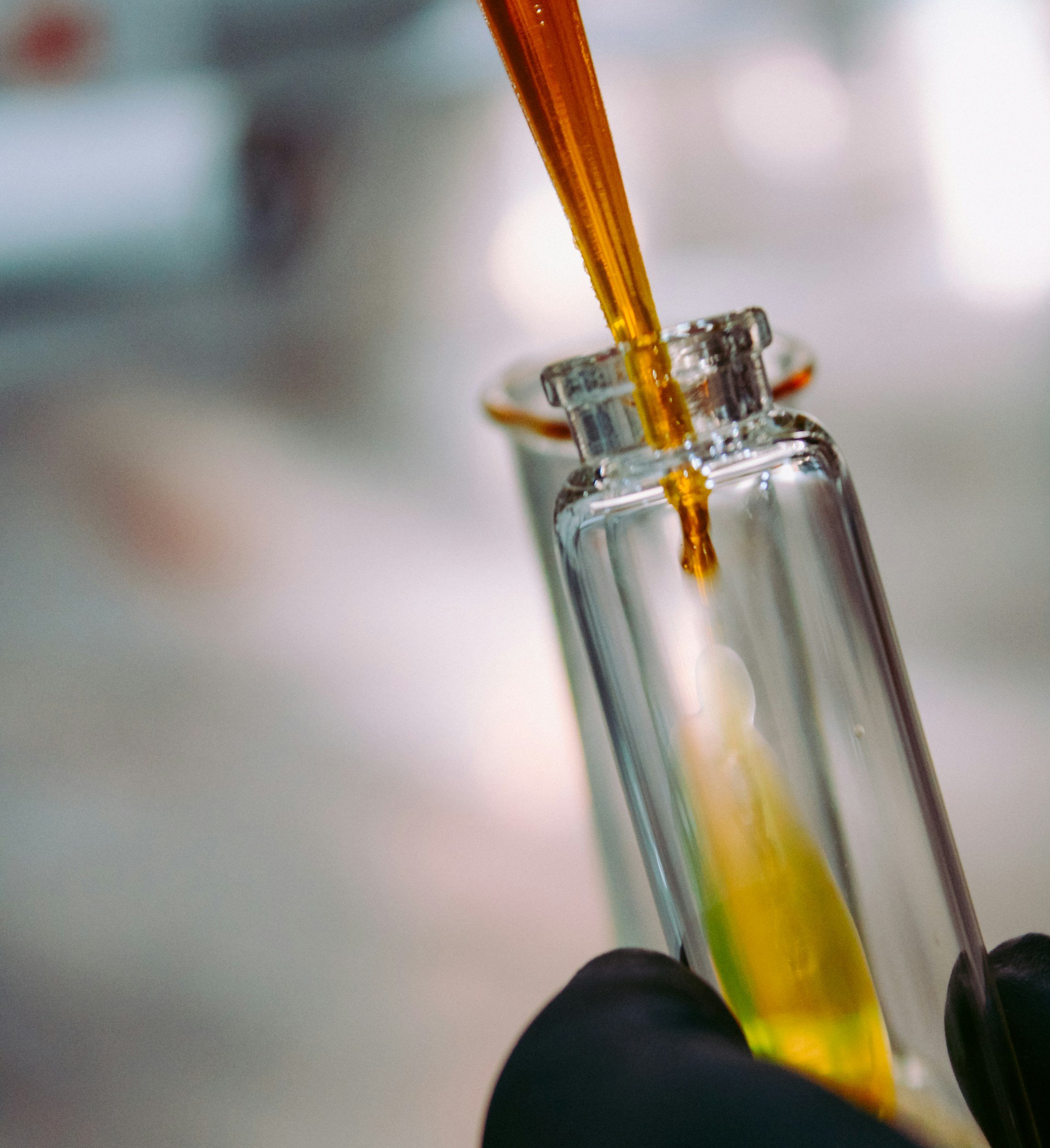 Close-up of a pipette transferring a yellow liquid into a small glass vial held by a gloved hand.