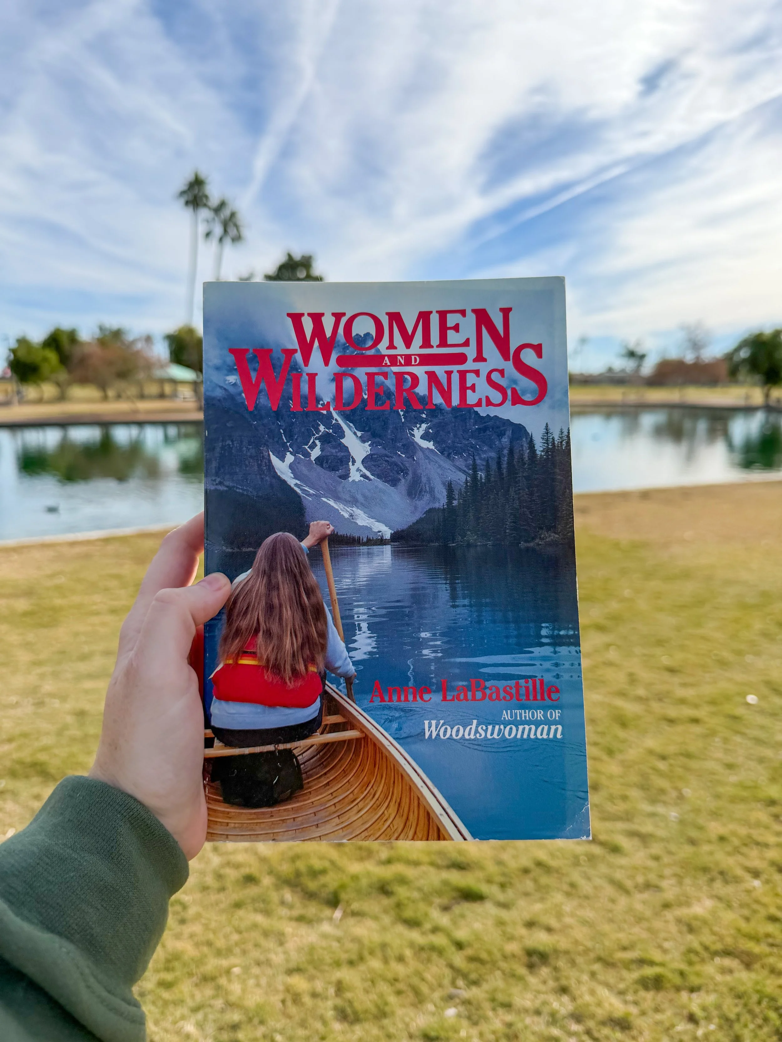 A person holding a book titled 'Women and Wilderness' by Anne LaBastille in front of a scenic outdoor park with a pond, trees, and a blue sky with wispy clouds.