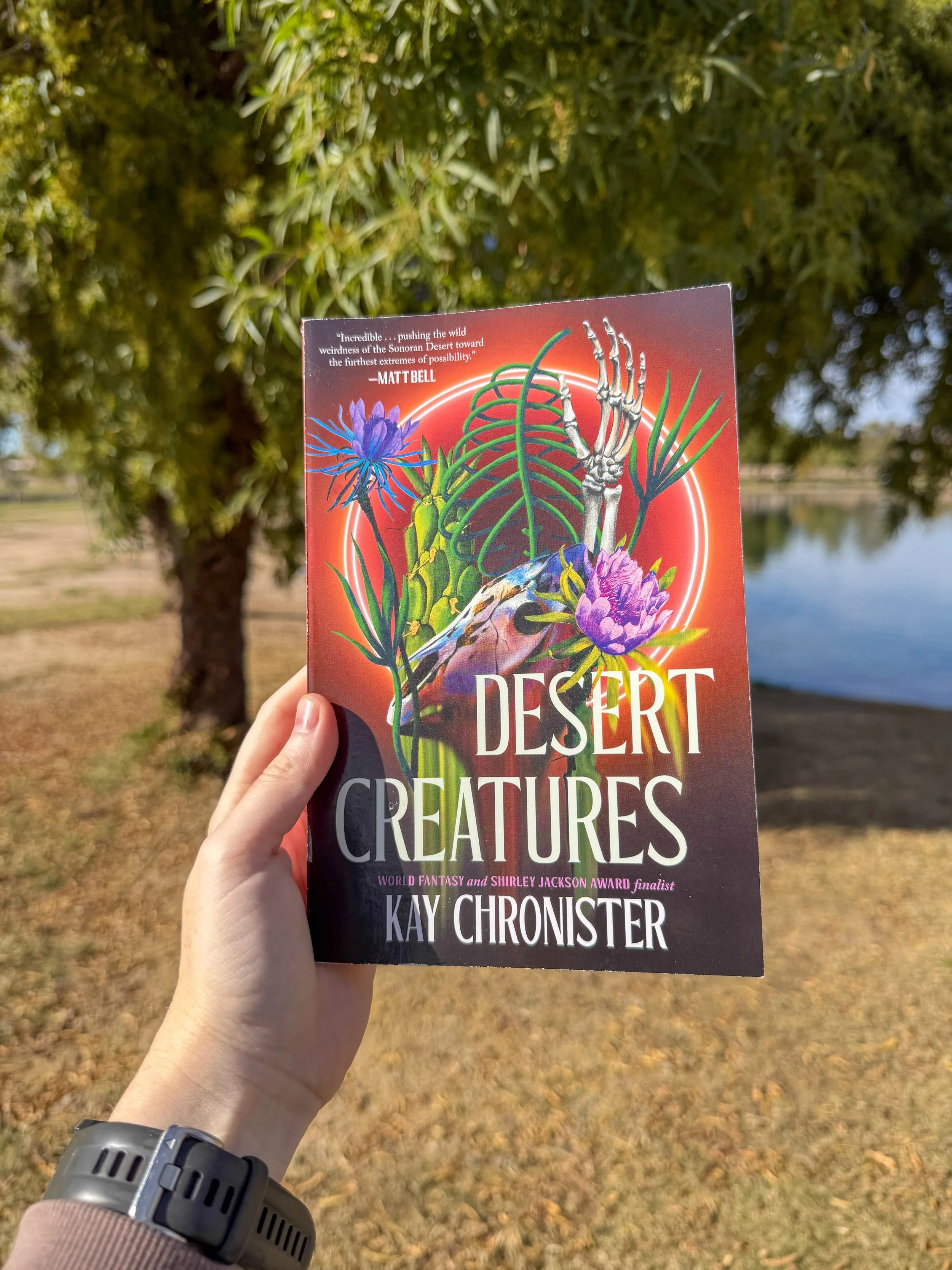 A person holding a book titled "Desert Creatures" by Kay Chronister outdoors near a body of water with trees and a grassy area in the background.