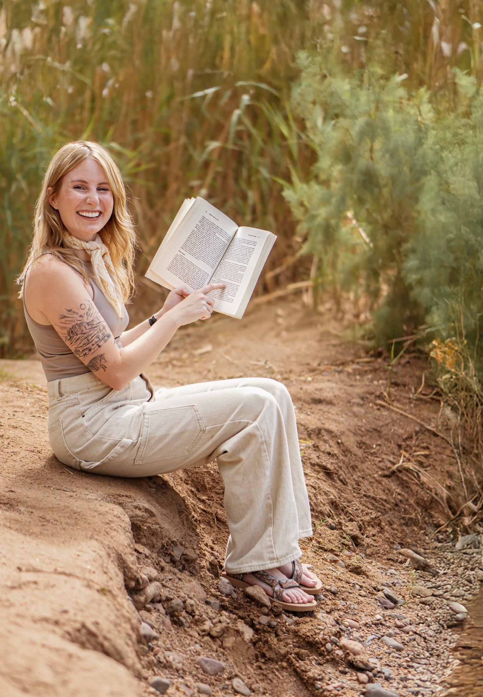 A young woman with blonde hair and tattoos, sitting on a dirt pathway near a body of water in a wooded area, smiling and reading a book.