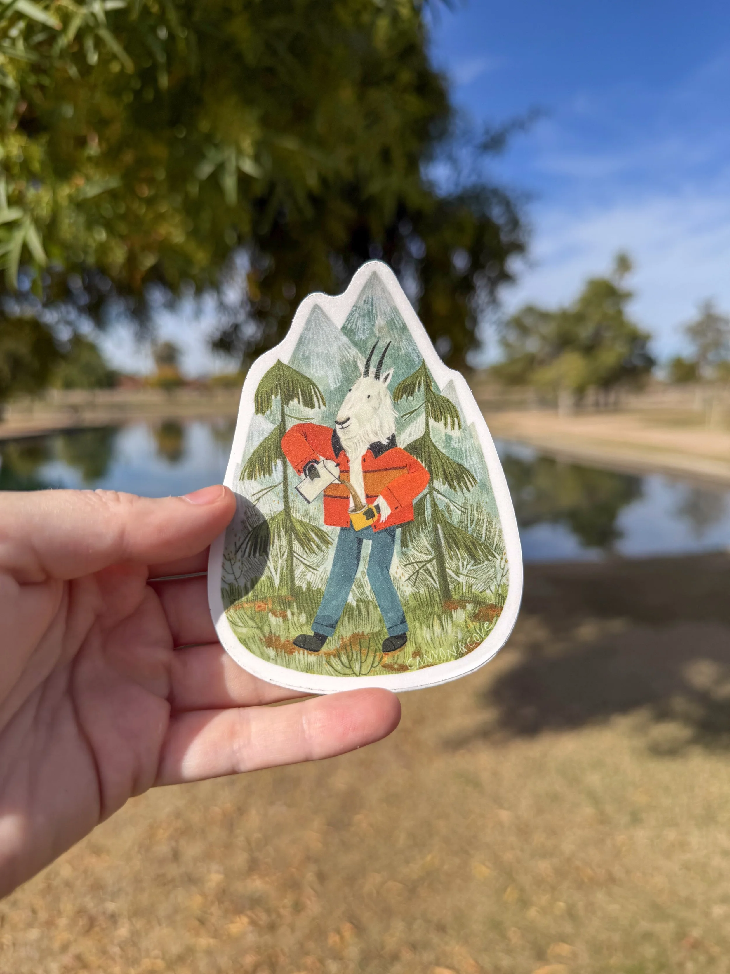 A person holding a sticker of an anthropomorphic goat with a camera, standing in a forest with mountains in the background.