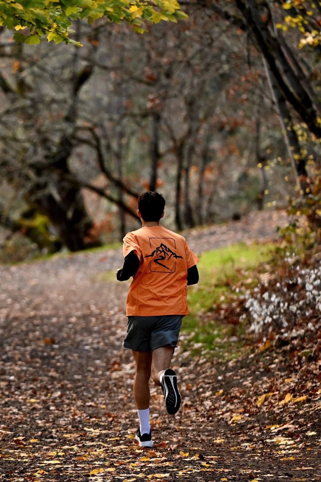 A man running on a dirt trail through a wooded area with autumn leaves on the ground and trees.