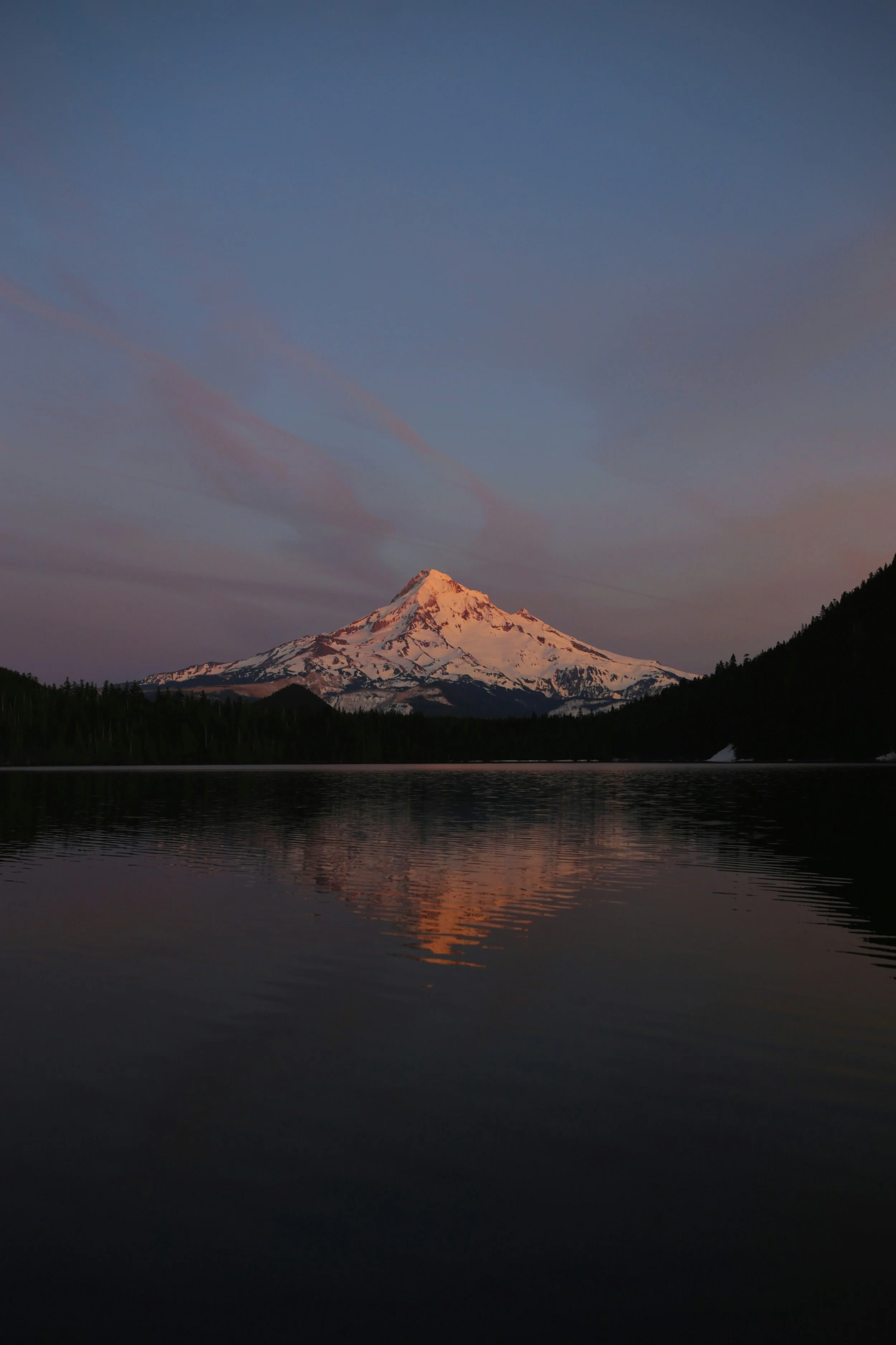 Snow-capped mountain reflecting in a calm lake at sunset or sunrise with pink and purple clouds in the sky.