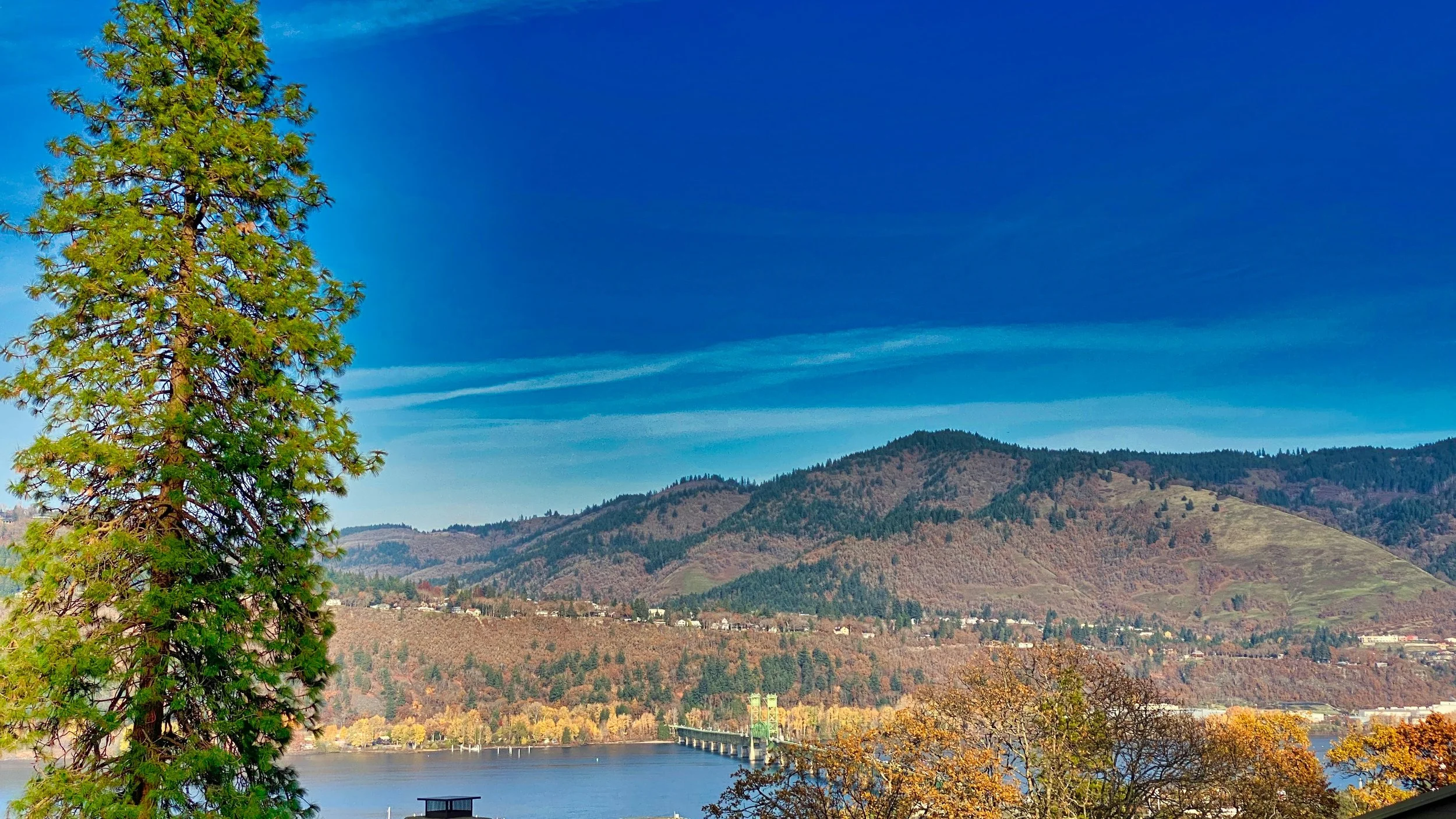 Scenic view of a lake with a bridge, surrounded by tree-covered hills and mountains under a bright blue sky with wispy clouds.