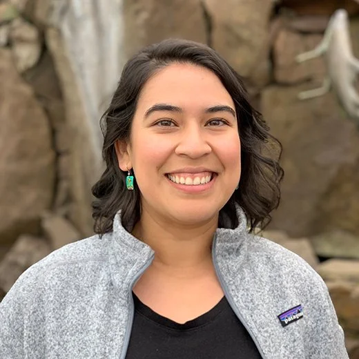 A smiling woman with dark wavy hair, wearing a gray jacket and turquoise earrings, standing outdoors near rocks.