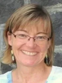 A smiling woman with short blonde hair and glasses, standing outdoors in front of a rocky background.
