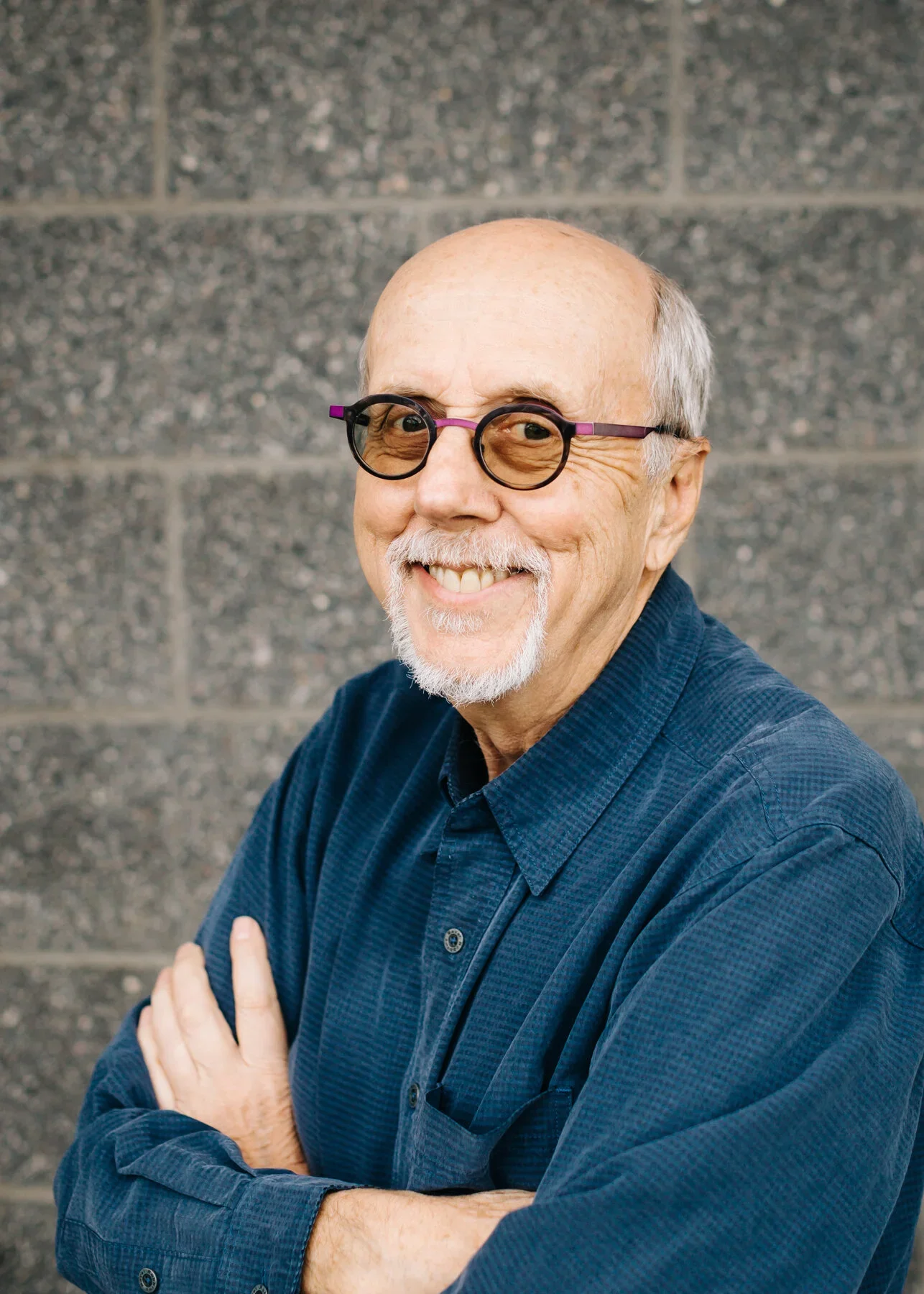 Smiling older man with glasses and white beard, wearing a dark blue shirt, standing against a gray brick wall.