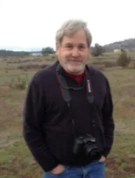 A man with gray hair and a beard smiling outdoors, dressed in a black jacket with a camera hanging around his neck.