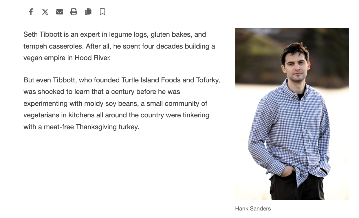 Photo of Hank Sanders standing outdoors near a body of water with trees in the background, wearing a blue checkered shirt and black pants, with his hands in his pockets.