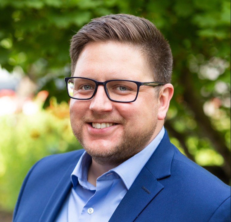 A man smiling outdoors, wearing glasses, a blue blazer, and a light blue shirt.