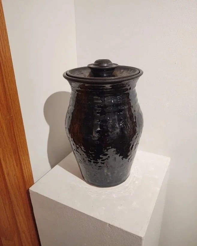 A black, shiny, textured ceramic vessel with a lid, placed on a white pedestal against a neutral wall.