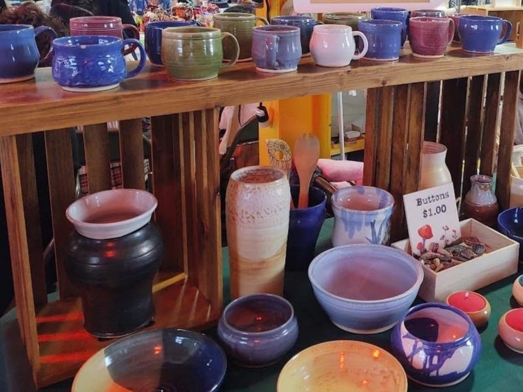 Display of handmade pottery mugs, bowls, and vases on wooden shelves at a craft fair.