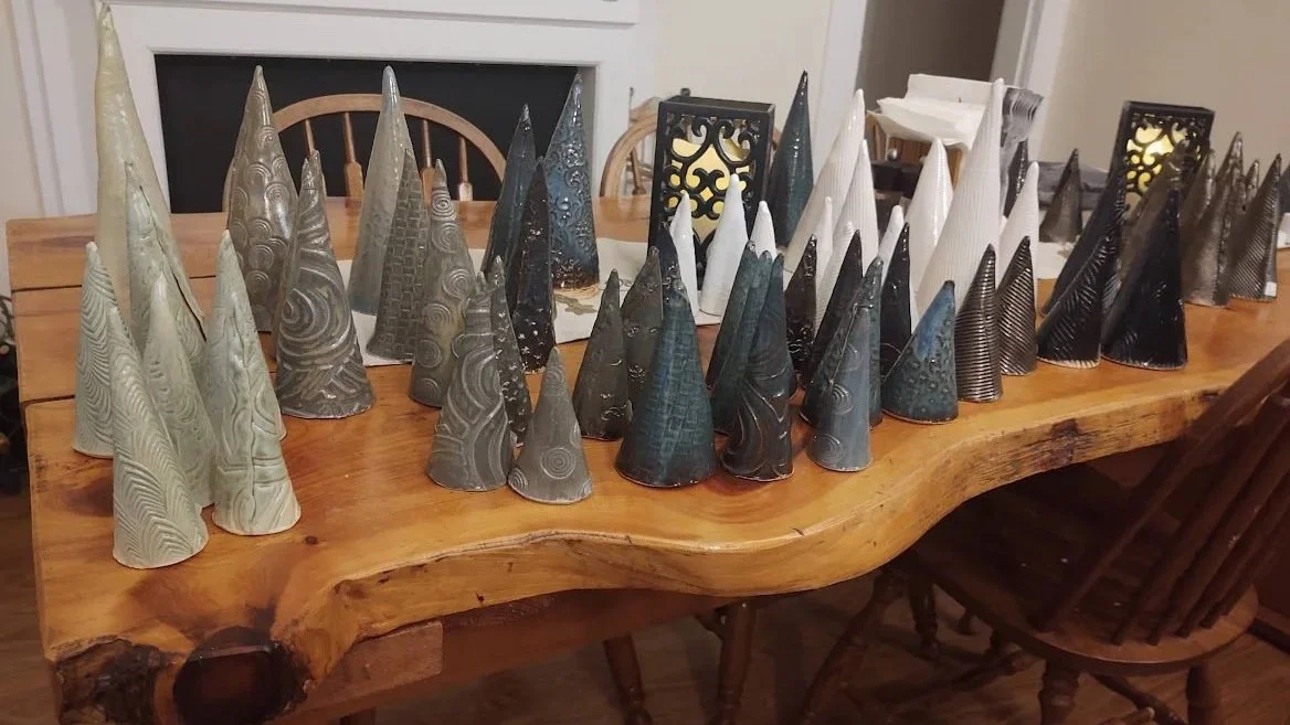 A wooden dining table decorated with assortments of ceramic Christmas trees in various sizes, shapes, and colors, with some in white, black, gray, and blue, arranged in a semi-circle.