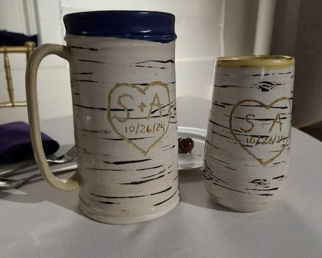 Two ceramic mugs with a white bark design and gold hearts with initials and date, placed on a table.