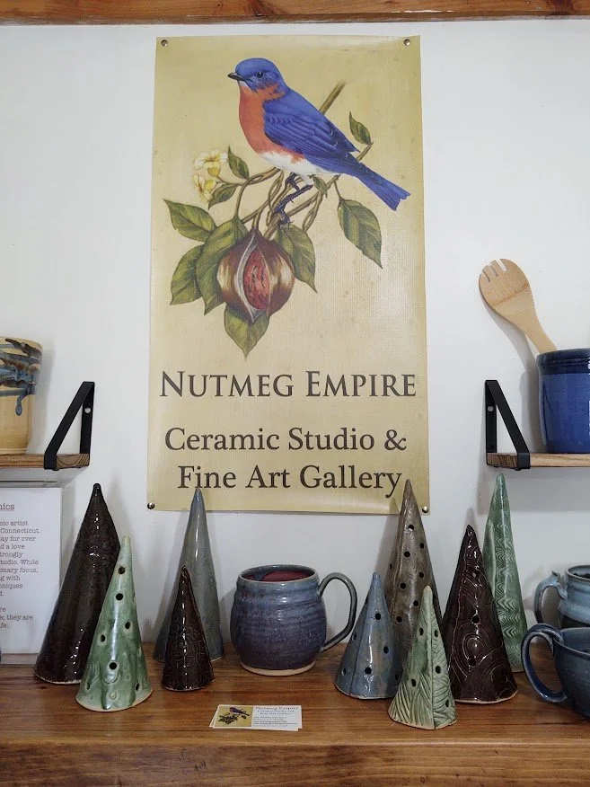 Display of ceramic cone-shaped art pieces in green, brown, and gray on a wooden surface in front of a sign for Nutmeg Empire Ceramic Studio & Fine Art Gallery, with a large poster of a bluebird perched on a branch.