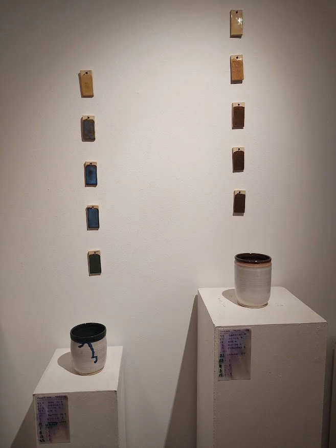 An art installation featuring two white pedestals with ceramic pots, and a wall behind with small rectangular color tiles arranged in two vertical lines.