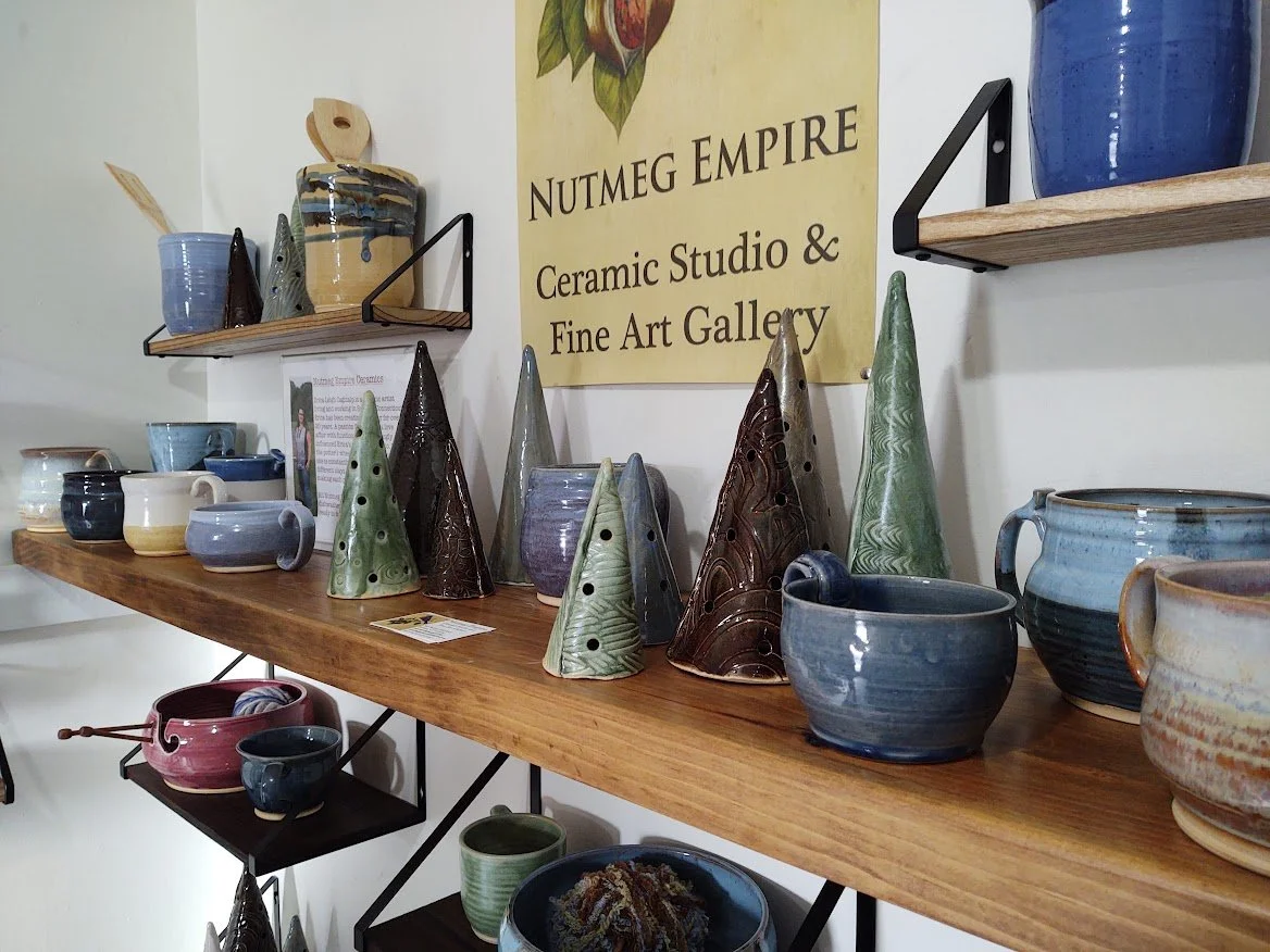 Display of ceramic pottery on shelves in a ceramic studio, with a sign reading 'Nutmeg Empire Ceramic Studio & Fine Art Gallery' in the background.